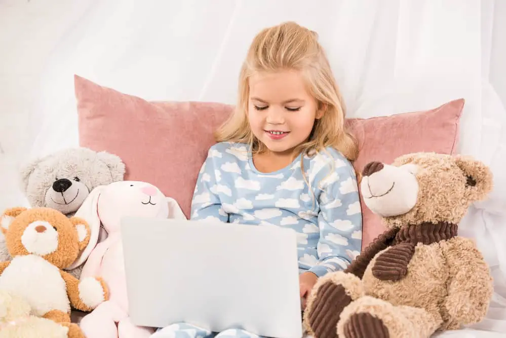 Little girl watching a laptop screen surrounded by teddy bears while her mother has quiet time with God.