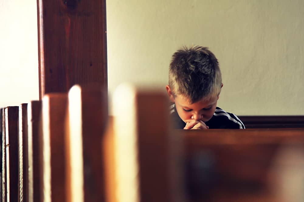 Young boy with his head bowed in prayer in a church pew. A penitent child saved and ready for baptism. 