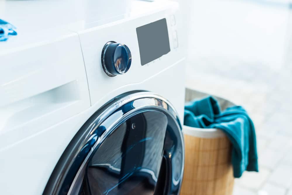 Sleek front loading washing machine in white, which you would have to use less with laundry hacks and tips. Bamboo laundry basket with blue clothing. 