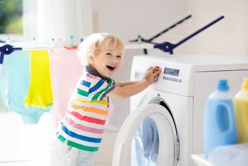 Little boy in rainbow striped shirt is helping with laundry in a bring laundry room.  