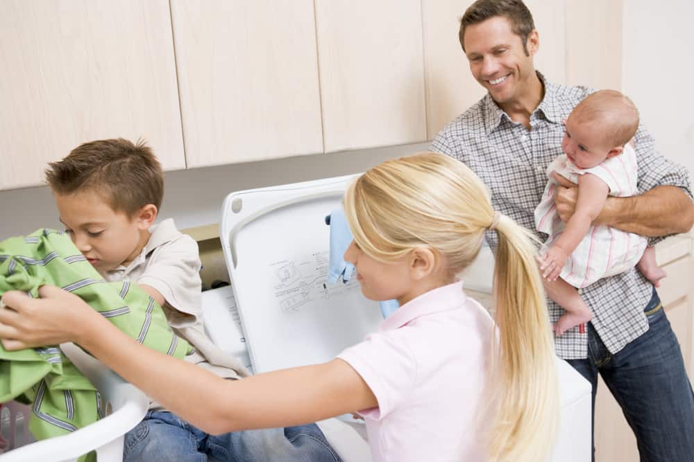 One of the best laundry hacks and tips is to get your family to help. Here is a picture of a family doing just that.  Dad is holding the baby while a brother and sister pull clothes out of the dryer and fold them. 