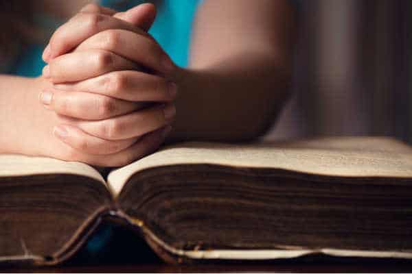 Woman in a turquoise blue shirt with her hands clasped in prayer over a very old Bible - example of pray without ceasing