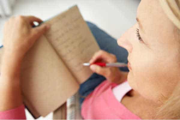 Woman in pink shirt is writing in a brown journal. Writing is one way of praying without ceasing. 