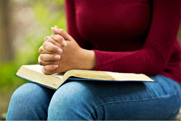 Woman in red/maroon top and jeans seated with a Bible open on her lap and her hands folded in prayer. 