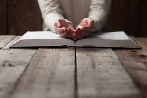 Gray wooden table.  Woman at the end with her hands open and turned up over her bible because she has a need for prayer for studying the Bible