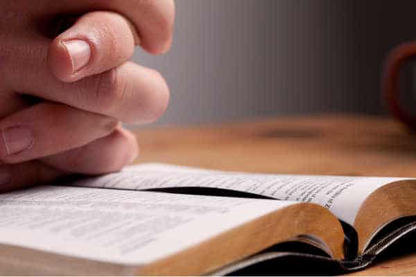 hands folded in prayer over a black open bible - living out the need for prayer for studying the Bible