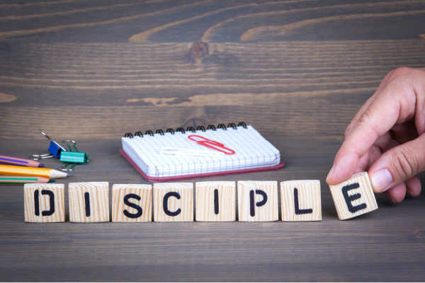 wood bakground.  small notebook with pink paperclips.  pencils and two binder clips.  Wooden block letters that spell out disciple.  A hand placing the last letter - e. - Discipling 