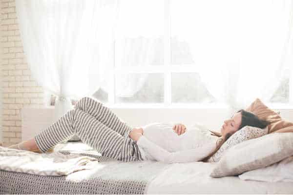 Pregnancy woman lying in bed.  She is wearing a white top and grey and white striped lounge pants.  She has her hands wrapped around her belly. Her eyes are downcast and she is meditating on pregnancy Bible verses.