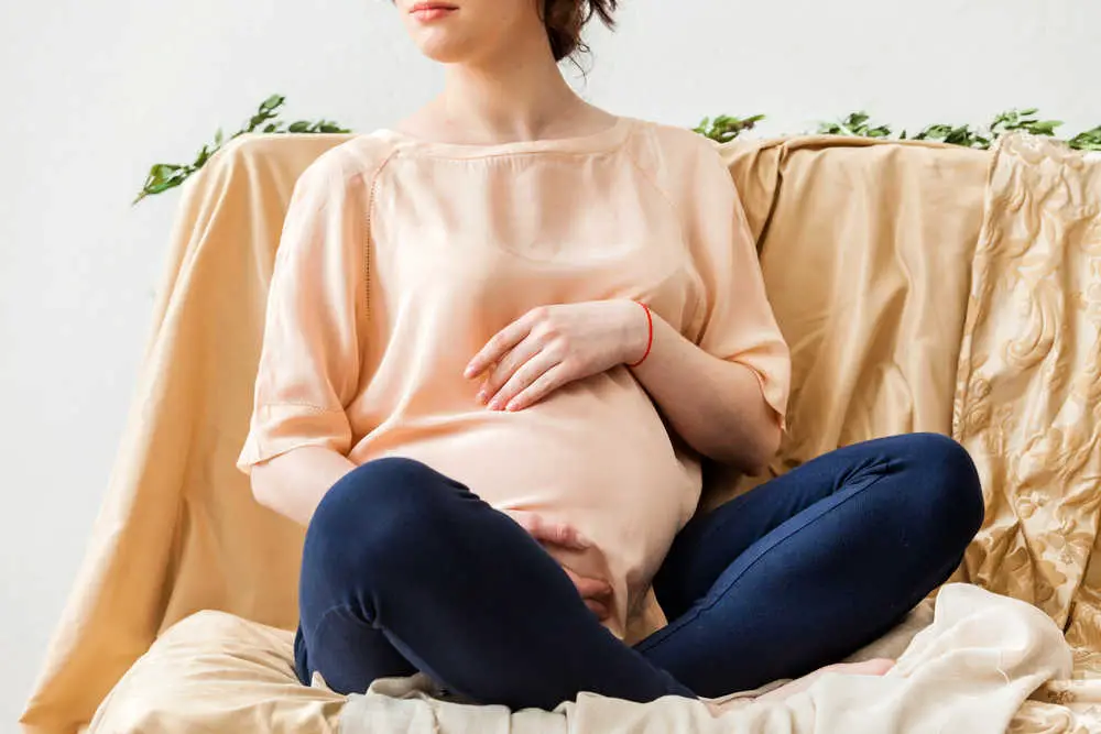 Pregnant woman in rose beige top and blue jeggings sitting against an artsy drapey cloth in beige with greenery across the top.  The woman is gently cradeling her belly and has a slightly anxious look on her face from pregnancy fears - she need pregnancy bible verses