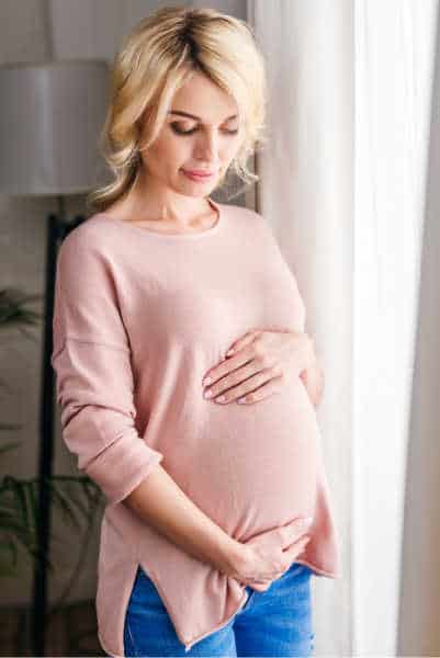 Woman standing cradeling her pregnant belly.  Soft pink, long sleeved shirt and jeans.  Soft white curtains are slightly behind her.  She is looking down with an anxious look on her face from pregnancy fears - she is meditating on pregnancy Bible verses