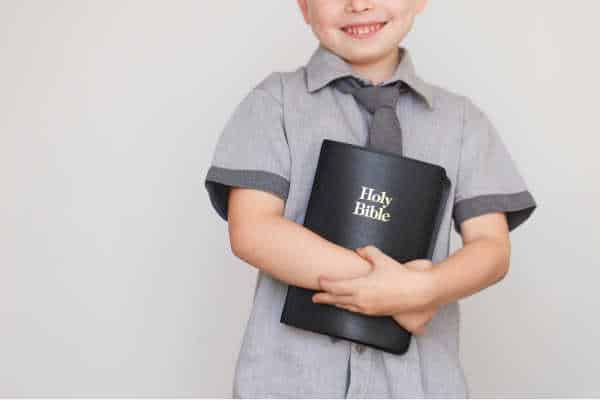 Little boy in gray shirt holding a black Bible on a gray background - he is heading to sunday school - the importance of sunday school