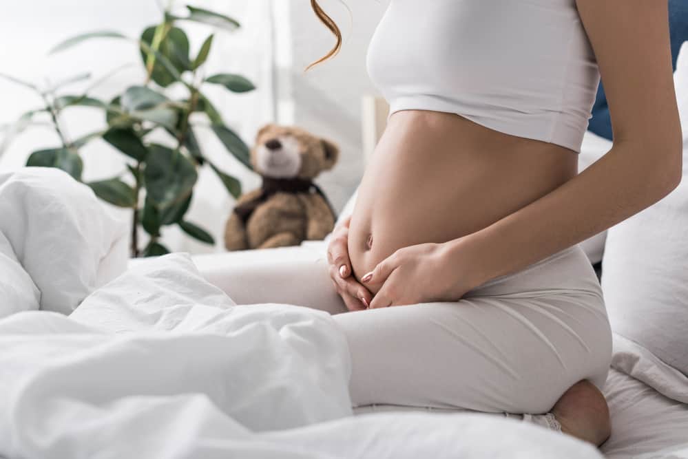 Calming white bedroom and bed.  Woman cradling her slightly enlarged belly - miscarriage fears and pregnancy fears while holding her belly