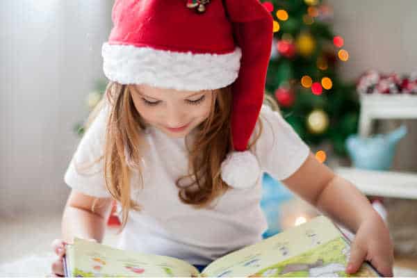 Little girl with Santa hat reading a book, because she is homeschooling during the holidays, with a Christmas tree in the background. 