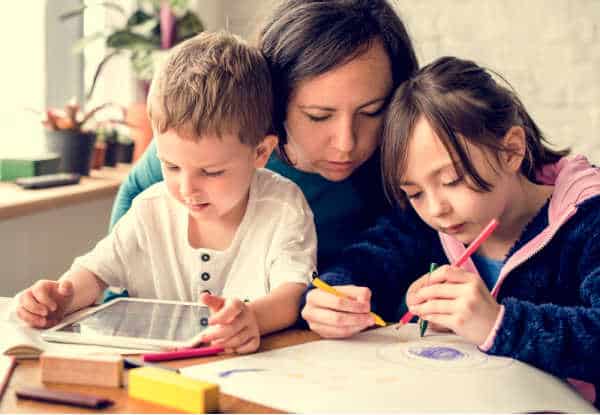 Homeschool mom is seated at a table with her two young children.  Her son is in her lab working on a tablet.  Her daughter is next to her drawing.  The mom is leaning over looking closely at what she is doing.  Homeschooling is more content, just like this picture, with bible verses for homeschool moms. 