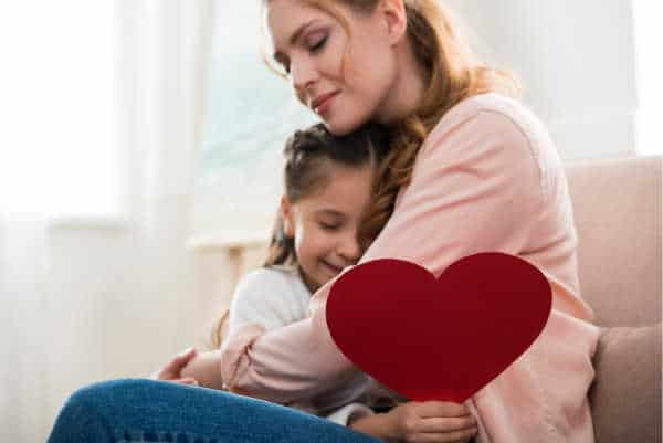 Mom sitting on a couch, hugging her daugther.  The daughter is holding a big red heart.  The mom has a serene look on her face - perhaps because she has bible verses for homeschool moms that she meditates on.