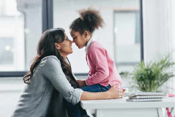 Little girl in a pink sweater and full pony tail sits on a table.  Her mom is sitting on a chair in front of her.  They are both smiling and are touching noses.  It is a picture of love and connection between a child and her homeschool mom. 