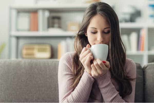 self-care for homeschool moms can look like a mom sitting on a couch savoring a cup of warm liquid in a white cup.  She is wearing a lavender thin sweater.  The couch is grey and their is a bookshelf in the background. 