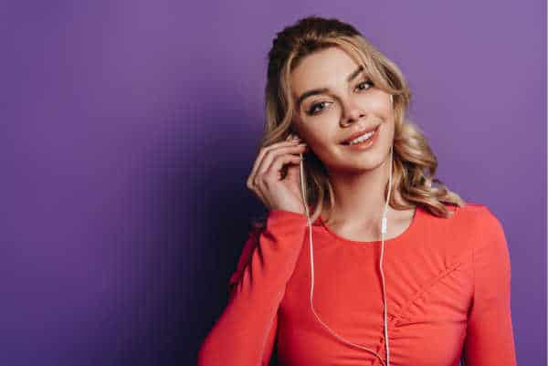 self-care for homeschool moms can look like listening to music, scripture, or an audiobook.  We don't know which one this mom is listening to yet.  She is wearing a coral long-sleeved top against a purple background.  She has blonde hair and brown eyes.  She is smiling at us as she puts in her second earbud. 