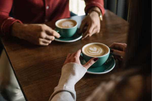 self-care for homeschool moms can look like going on a date - without your kids.  Here we see a mom and dad having coffee together.  We see just the hands and the coffee, green cups with hearts in the froth. 