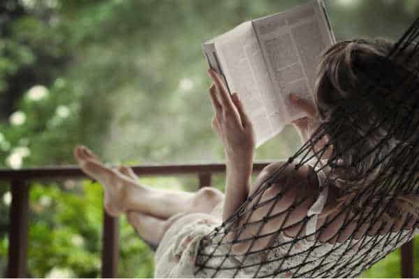 Woman lying in a hammock in a garden and enjoying a book reading, this is also what self-care for homeschool moms can look like.  She is wearing a dress and is barefooted.  It looks like she is reading the Bible. There is green beyond her on the porch. 