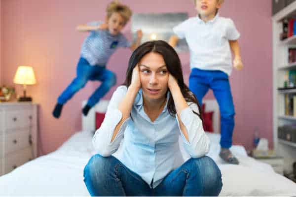 Mom wondering how long her quiet time needs to be because her kids are up and already bouncing on the bed.  She is sitting in the foreground with her hands over her ears while he two boys jump on the bed behind her. 