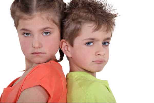 Little girl in an orange shirt with her back to a little boy in green shirt.  Both have grumpy looks on their face because mom and dad need to parent a bossy child - big sister!