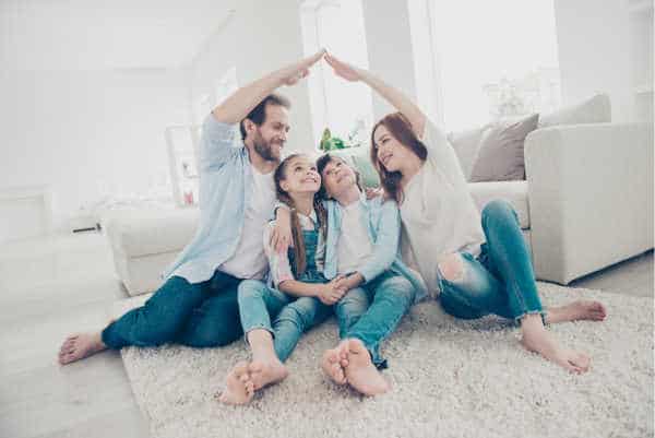 Family of four sitting on the floor in a bright, airy living room.  Mom and Dad have their arms raised over the kids and touching like a roof - when you parent a bossy child you need to give them submission to the authority structure of a family.