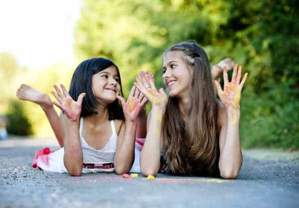 We parent a bossy child to be better siblings and friends.  Here is a big sister and a little sister with hands covered in chalk.  They are lying on the ground drawing together.  At the moment that are looking at each other and smiling as they show off their chalk covered hands. 