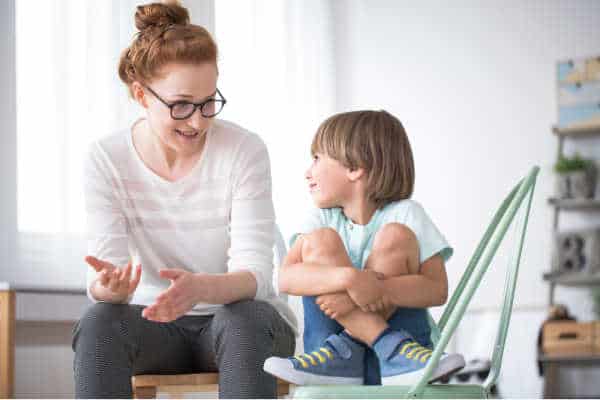 You parent a bossy child like the woman pictured here.  She is firm but open in talking to a little boy.  He is sitting with his legs crossed on a chair as if he is in timeout.  She is leaning forward as she sits to connect with him and coach him on his behavior. 