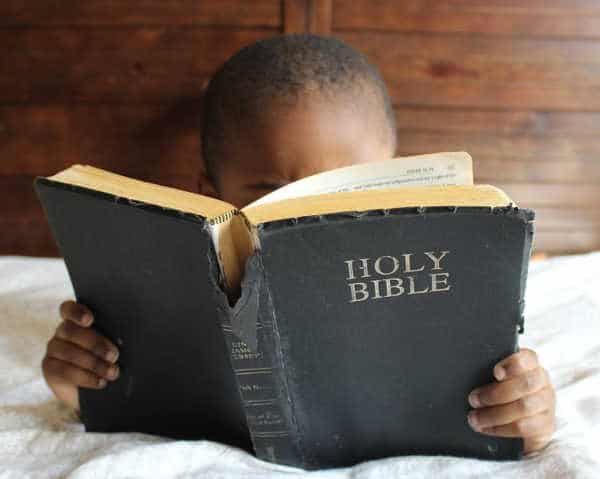 Little African-American boy reading a well-worn copy of the Bible.  He face is mostly hidden by it.  He is reading the Bible and wants to memorize the Books of the Bible.