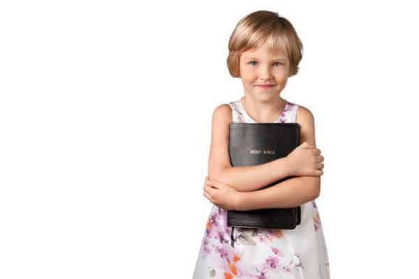 Little blonde haired girl with a pixie cut and dressed in a sleeveless white floral dress is squeezing a black Bible in front of her in a hug.  She is excited because is working hard to memorize the Books of the Bible.