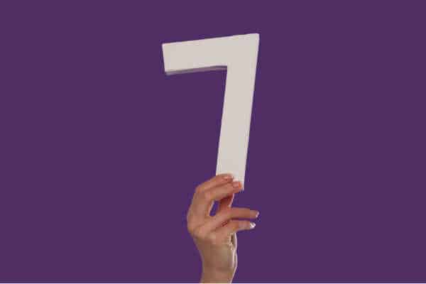 female hand holding up the number 7 against a purple background - It is helpful to memorizeMemorize the Books of the Bible in sets of 7