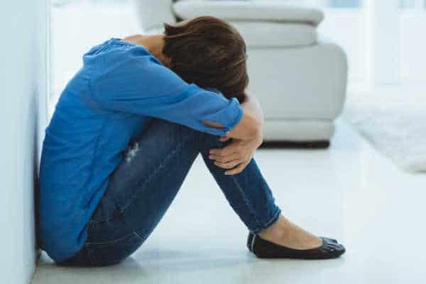 Mom overcome with mom guilt with is sitting on the floor with her head against her knees, and her arms wrapped around her knees.  She is dressed in a blue top and blue jeans. 