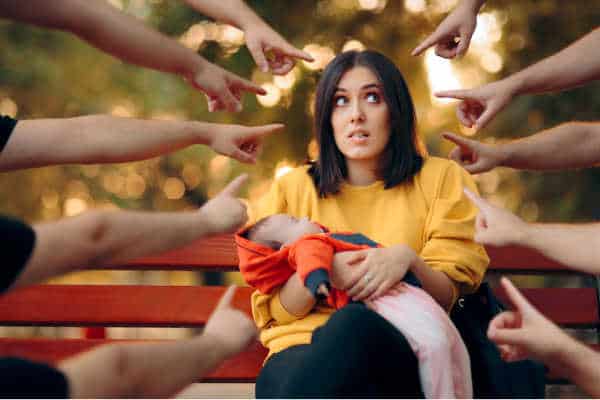 A woman sitting on a bench holding her baby filled with mom guilt because all kinds of hands are pointing at her. 