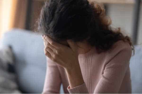 Mom with dark, long hair and a pink sweater.  She is struggling with mom guilt and is sitting on the couch with her face buried in her hands. 