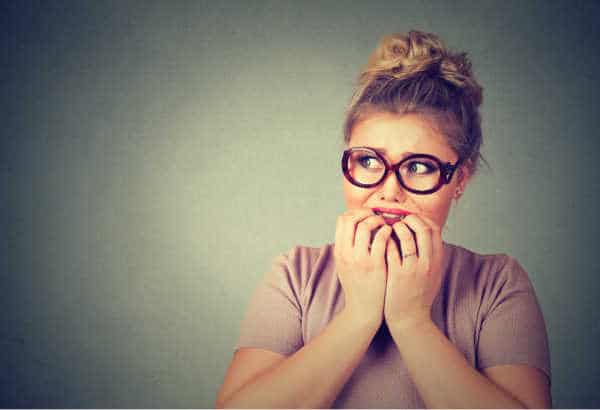 Gray background.  Mom in light mauve tshirt with hair in a mom bun.  She has thick glasses and she is biting her fingernails.  Her expression is full of mom guilt and anxiety. 