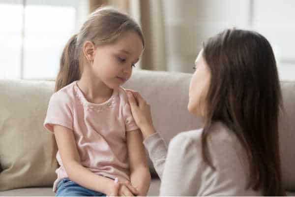 Mom filled with mom guilt is prompted to apologize to her daughter.  The young daughter is sittng on the couch with a sad look.  The mom's face is turned from us and to the little girl.  She has her hand on the girl's shoulder. 