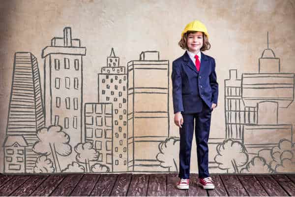 Young boy in a business suit, red keds, and a hard hat.  There is a black and white drawing of a skyline in the background.  It is a smiling youn boy - prepare your homeschooler to be an entreprenuer
