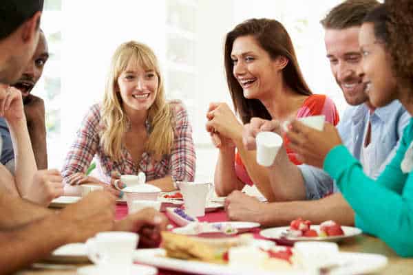 A Homemaker is hospitable like the woman here who has a diverse group of friends gathering closely around a table talking, laughing, and eating. 