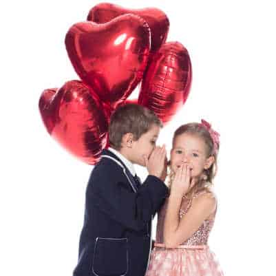 Young boy with a bouquet of heart balloons whispers into the ear of a young girl on St. Valentine's Day. 