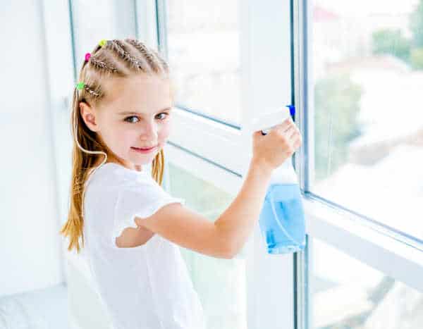 Little girl in a white blouse cleaning a window with blue cleaner on behalf of an unappreciated mom